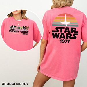 May include: A pink t-shirt with a Disney Crew graphic on the front and a Star Wars design on the back. The Star Wars design includes a sunset, a spaceship, and the text "STAR WARS 1977".