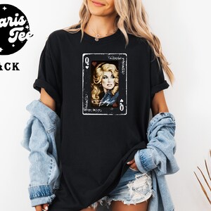 May include: A black t-shirt featuring a graphic print of a playing card with the letter Q and a heart symbol. The card displays a portrait of a blonde person with wavy hair, wearing a denim jacket, with the name Dolly written in white script below.