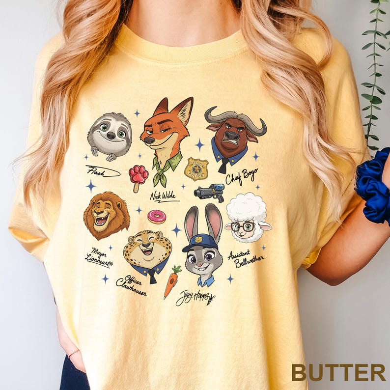 K&ouml;nnte beinhalten: Gelbes T-Shirt mit Cartoon-Illustrationen von Zootopia-Charakteren, darunter Flash, Nick Wilde und Judy Hopps. Das Wort "BUTTER" ist unten rechts auf dem T-Shirt aufgedruckt.