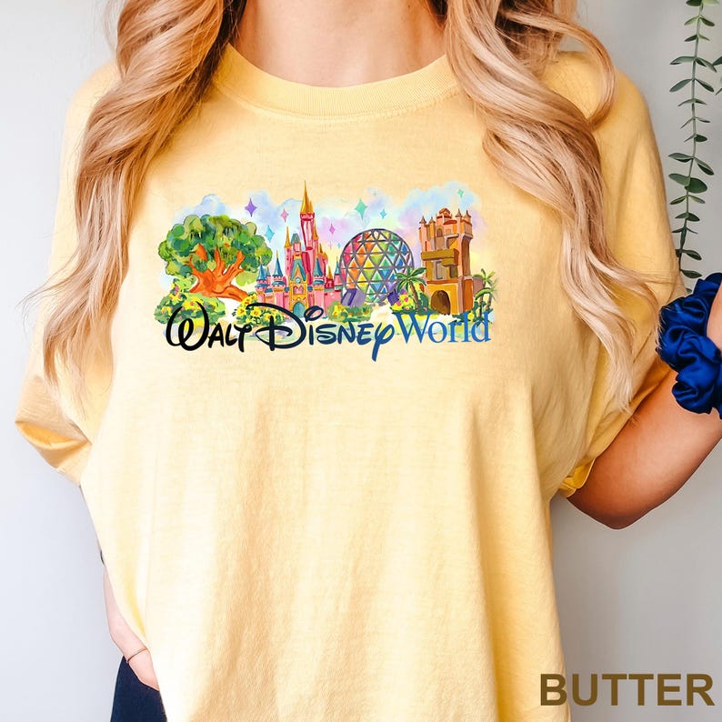May include: A butter yellow t-shirt featuring a colorful graphic print of Walt Disney World landmarks, including a large tree, a pink castle, a geodesic dome, and a tower, with the text Walt Disney World written in a dark blue script font.