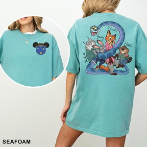 May include: Seafoam green t-shirt featuring a cartoon graphic on the front and back. The front graphic is a blue circle with a mouse-eared design. The back graphic features cartoon characters and the names Judy, Nick, and Nibbles.