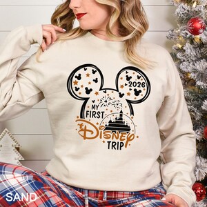 May include: Sand-colored sweatshirt with a black and brown Mickey Mouse head design. The design includes the text "My First Disney Trip" and the year "2026". The sweatshirt is long-sleeved.