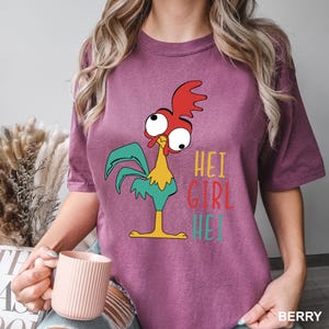 May include: Berry-colored t-shirt featuring a cartoon rooster with red comb, teal wings, and yellow body. The text "HEI GIRL HEI" is printed in yellow, red, and teal. The shirt is a comfortable fit.