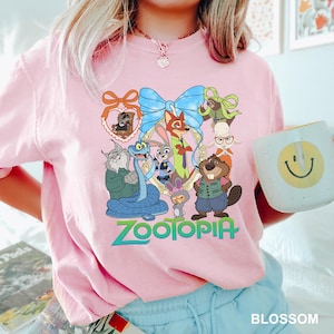 K&ouml;nnte beinhalten: Ein hellrosa T-Shirt mit einer farbenfrohen Grafik von Charakteren aus dem Film "Zoomania". Das Design umfasst eine blaue Schleife, das Wort "Zootopia" in Gr&uuml;n und verschiedene Tierfiguren. Eine wei&szlig;e Tasse mit einem gelben Smiley ist ebenfalls sichtbar.