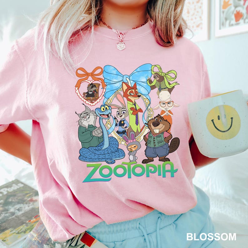 K&ouml;nnte beinhalten: Ein hellrosa T-Shirt mit einer farbenfrohen Grafik von Charakteren aus dem Film "Zoomania". Das Design umfasst eine blaue Schleife, das Wort "Zootopia" in Gr&uuml;n und verschiedene Tierfiguren. Eine wei&szlig;e Tasse mit einem gelben Smiley ist ebenfalls sichtbar.