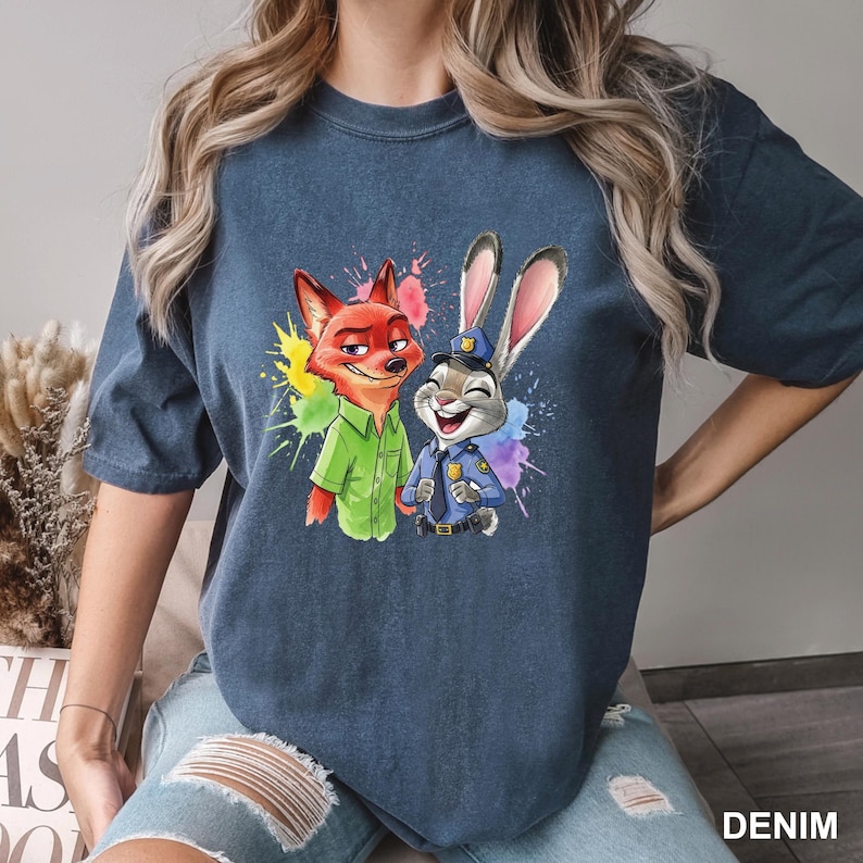 May include: Denim blue t-shirt featuring a colorful graphic of Nick Wilde and Judy Hopps from Zootopia. Nick is wearing a green shirt, and Judy is in a police uniform. The word "DENIM" is printed on the bottom right.