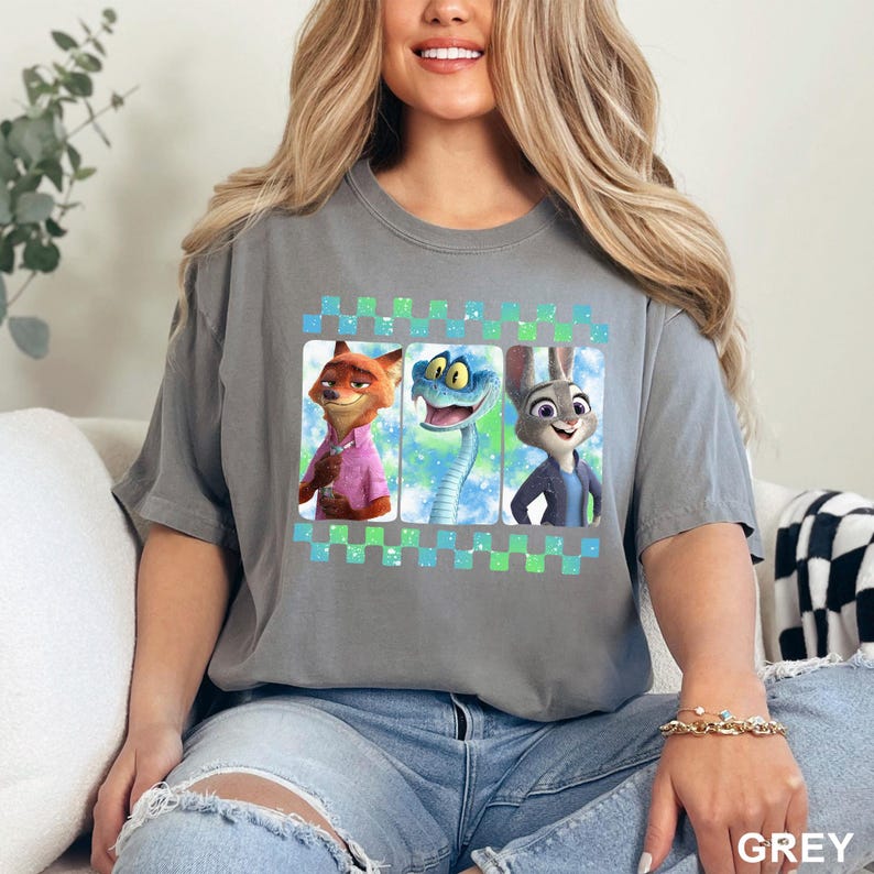 May include: A grey t-shirt featuring three cartoon characters in rectangular frames. The characters include a fox in a pink shirt, a snake, and a rabbit. The frames are set against a blue and green checkered pattern.