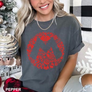 May include: A dark gray t-shirt with a red graphic design. The design is a circular arrangement of various shapes and symbols, with a mountain-like shape in the center. The word "PEPPER" is at the bottom of the image.
