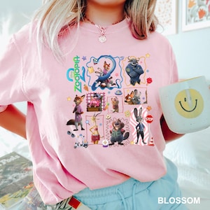 May include: A light pink t-shirt featuring a colorful graphic print of various Zootopia characters, including Nick Wilde and Judy Hopps, arranged in a collage style with the Zootopia logo, small icons, and a stop sign, all printed on the front chest.