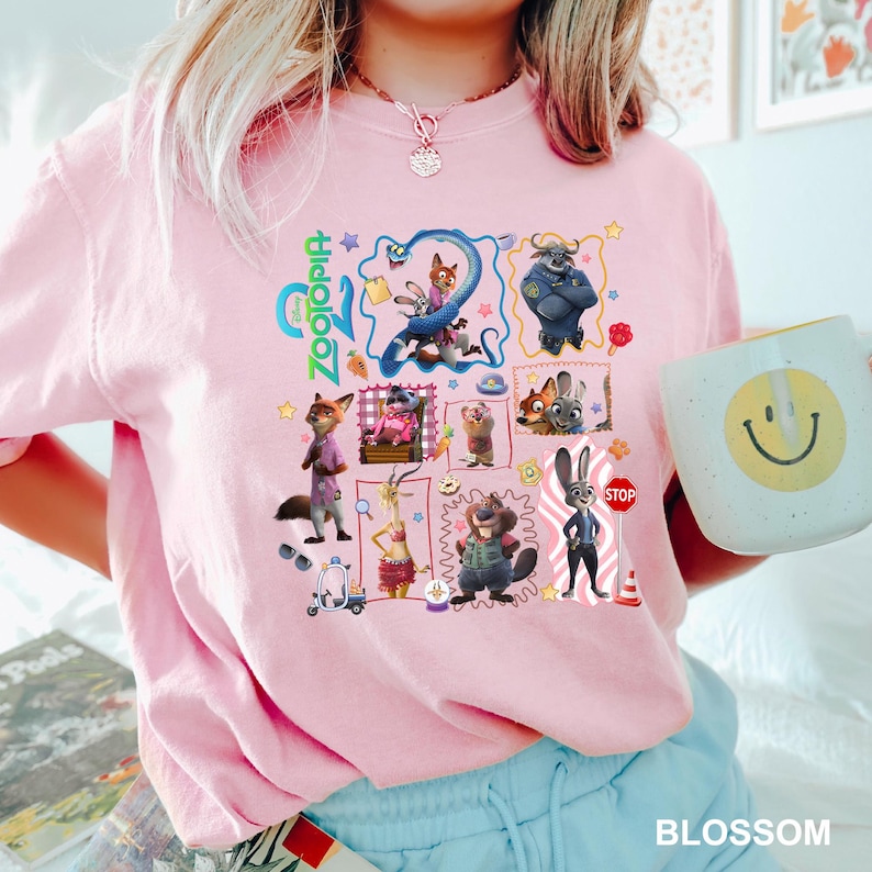 May include: A light pink t-shirt featuring a colorful graphic print of various Zootopia characters, including Nick Wilde and Judy Hopps, arranged in a collage style with the Zootopia logo, small icons, and a stop sign, all printed on the front chest.