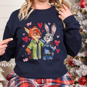 K&ouml;nnte beinhalten: Marineblaues Sweatshirt mit einer Aquarellillustration von Nick Wilde und Judy Hopps aus Zoomania, umgeben von roten Herzen. Der Text "NAVY" ist auf der linken Seite des Sweatshirts aufgedruckt.