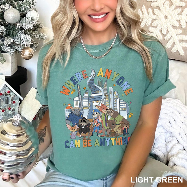 May include: Light green t-shirt with the text "WHERE ANYONE CAN BE ANYTHING" in a colorful, retro font. The graphic features cartoon animals and city buildings. The shirt is a comfortable, casual style.