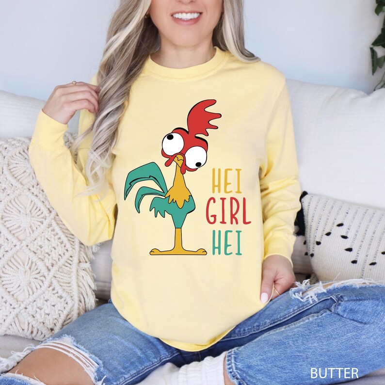 May include: A light yellow long-sleeve shirt featuring a cartoon rooster with red, green, yellow, and blue accents. The shirt has the text "HEI GIRL HEI" in colorful letters. The shirt is made of a soft material.