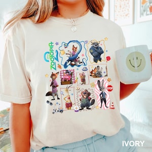 May include: An ivory t-shirt featuring a colorful Zootopia graphic print with various characters including Judy Hopps and Nick Wilde, alongside small icons like a stop sign, a scooter, and stars, arranged in a collage style on the front.