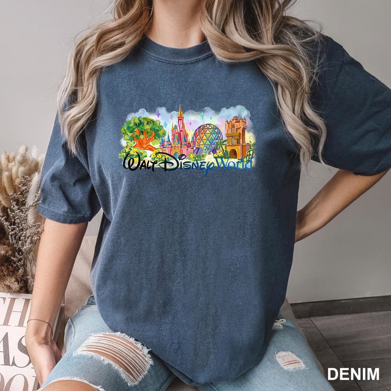 May include: A dark blue short-sleeve t-shirt featuring a colorful graphic print of Walt Disney World landmarks, including a castle, a geodesic sphere, and a tree. The text Walt Disney World is printed in black script below the colorful illustration.