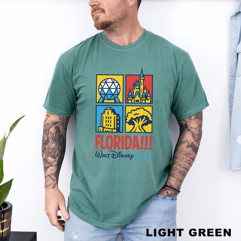 Może przedstawiać: Jasnozielona koszulka z kolorową grafiką przedstawiającą cztery kwadraty z kultowymi zabytkami Florydy. Tekst "FLORIDA!!!" i "Walt Disney World" są wydrukowane poniżej. Koszulka jest wykonana z miękkiego materiału.