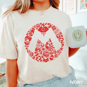 May include: Ivory-colored t-shirt featuring a red graphic design of a video game character. The design is a circular arrangement of game-related icons. A white mug with a smiley face is held in the image.