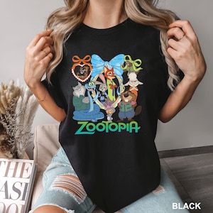 K&ouml;nnte beinhalten: Schwarzes T-Shirt mit einer farbenfrohen Grafik von Zootopia-Charakteren, darunter Judy Hopps und Nick Wilde, in einem dekorativen Rahmen. Das Wort "Zootopia" ist in Gr&uuml;n und Blau dargestellt. Das T-Shirt ist schwarz.