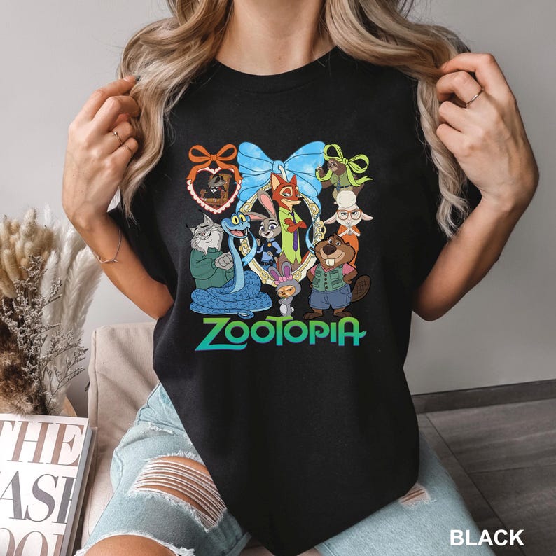 K&ouml;nnte beinhalten: Schwarzes T-Shirt mit einer farbenfrohen Grafik von Zootopia-Charakteren, darunter Judy Hopps und Nick Wilde, in einem dekorativen Rahmen. Das Wort "Zootopia" ist in Gr&uuml;n und Blau dargestellt. Das T-Shirt ist schwarz.