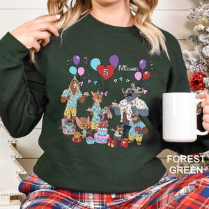 May include: Forest green sweatshirt featuring a colorful cartoon birthday party scene with animal characters, balloons, and a cake. The number 5 and the name "Michael" are also displayed. The sweatshirt is paired with plaid pajama pants.