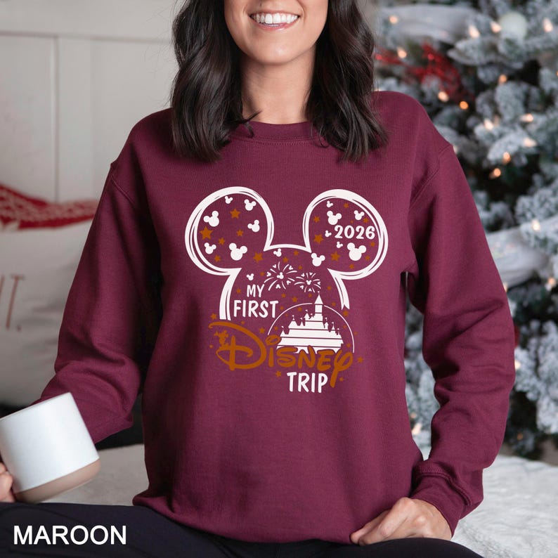 May include: Maroon sweatshirt with a white Mickey Mouse head design. The design includes the text "My First Disney Trip" and the year "2026". The sweatshirt is a solid maroon color.