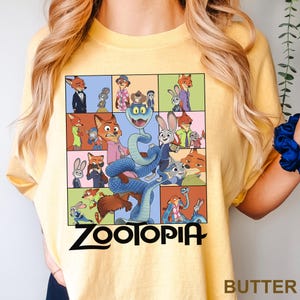 May include: Yellow t-shirt featuring a colorful graphic from the animated movie Zootopia. The design includes various characters like Nick Wilde, Judy Hopps, and a blue snake, with the word "Zootopia" in bold letters. The word "BUTTER" is printed at the bottom right.