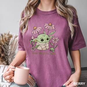 Puede incluir: Camiseta color bayas con un gr&aacute;fico de un personaje alien&iacute;gena verde rodeado de flores rosas y blancas y una peque&ntilde;a rana. El texto "ENJOY THE LITTLE THINGS" est&aacute; impreso debajo. La camiseta es de manga corta y cuello redondo.
