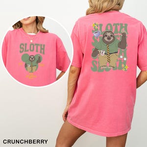 May include: A pink t-shirt featuring a sloth cartoon design. The front displays "SLOTH" above a sloth character with a coffee cup. The back has a larger sloth graphic with "SLOTH" and "SLOTH" in green. The shirt is a comfortable fit.