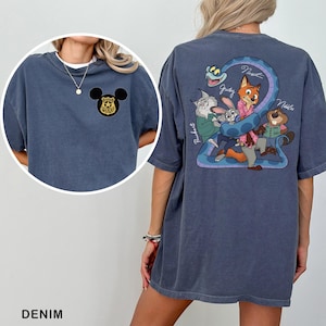 May include: Denim blue t-shirt featuring a Mickey Mouse police badge on the front and a cartoon graphic on the back. The graphic includes characters like Judy, Nick, and others, with the names of the characters written on the shirt.