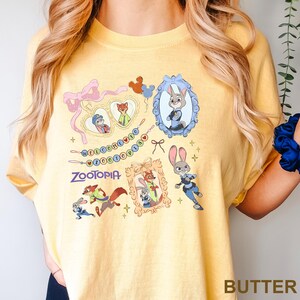 Op de afbeelding: Boterkleurig T-shirt met Zootopia-personages en afbeeldingen. Het ontwerp bevat ingelijste afbeeldingen van Judy Hopps en Nick Wilde, samen met het woord "Zootopia". Extra decoratieve elementen zijn harten, strikken en sterren.