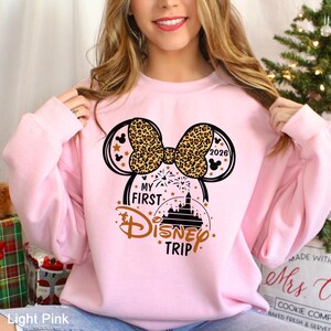 May include: Light pink sweatshirt with a graphic featuring a leopard print bow, Mickey Mouse ears, and the text "My First Disney Trip 2026." The design includes stars and a castle silhouette.