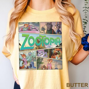 K&ouml;nnte beinhalten: Ein blassgelbes T-Shirt mit einem Grafikdruck von Zootopia-Figuren im Comic-Stil. Das Design enth&auml;lt mehrere Szenen mit Nick Wilde und Judy Hopps, wobei das Wort Zootopia in gro&szlig;en gr&uuml;nen Buchstaben prominent dargestellt ist.