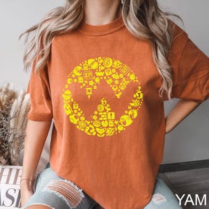 May include: Burnt orange t-shirt featuring a yellow graphic design. The design is a stylized "W" shape composed of various video game icons. The shirt has short sleeves and a relaxed fit. The text "YAM" is visible in the bottom right corner.