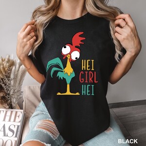 May include: Black t-shirt featuring a cartoon rooster with red, yellow, and teal accents. The text "HEI GIRL HEI" is printed in colorful letters. The shirt is a crew neck and has short sleeves.