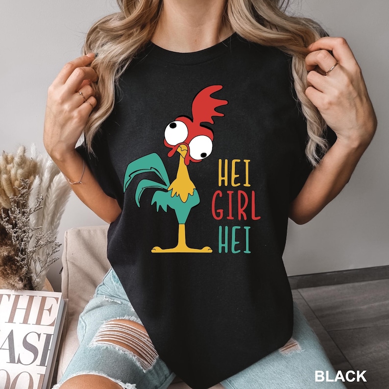 May include: Black t-shirt featuring a cartoon rooster with red, yellow, and teal accents. The text "HEI GIRL HEI" is printed in colorful letters. The shirt is a crew neck and has short sleeves.
