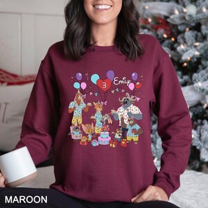 May include: Maroon sweatshirt featuring a colorful cartoon birthday party scene with animal characters, balloons, and a cake. The name "Emily" and the number "3" are also displayed. The word "MAROON" is printed at the bottom.
