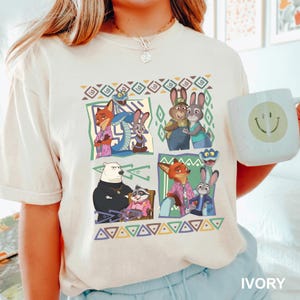 K&ouml;nnte beinhalten: Elfenbeinfarbenes T-Shirt mit einer farbenfrohen Grafik von animierten Tierfiguren in verschiedenen Szenen. Das Design umfasst F&uuml;chse, Kaninchen und andere Tiere in einem geometrischen Muster. Eine wei&szlig;e Tasse mit einem Smiley wird im Bild gehalten.