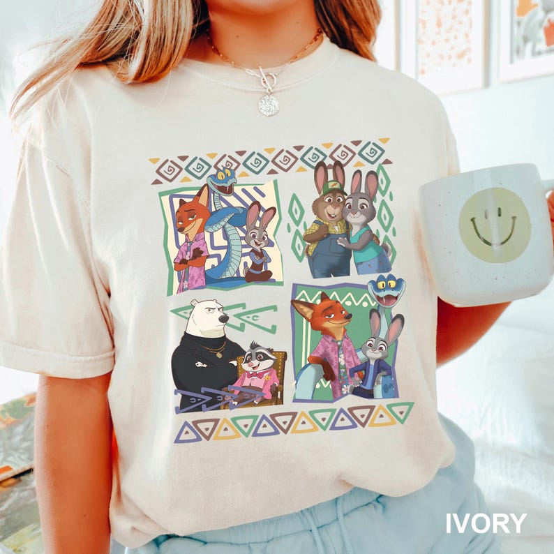 K&ouml;nnte beinhalten: Elfenbeinfarbenes T-Shirt mit einer farbenfrohen Grafik von animierten Tierfiguren in verschiedenen Szenen. Das Design umfasst F&uuml;chse, Kaninchen und andere Tiere in einem geometrischen Muster. Eine wei&szlig;e Tasse mit einem Smiley wird im Bild gehalten.