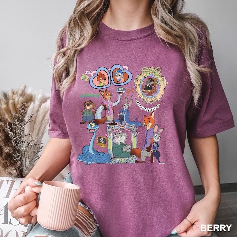 May include: Berry-coloured t-shirt with a cartoon graphic of animal characters, including framed portraits and the word "Zootopia". The shirt is made of a soft material.
