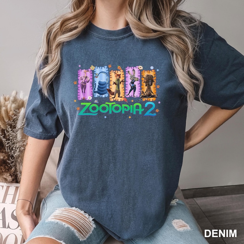 K&ouml;nnte beinhalten: Ein dunkelblaues T-Shirt mit einem farbenfrohen Grafikdesign aus dem Film "Zootopia 2". Das Design zeigt verschiedene Charaktere und den Filmtitel in einem hellen, ansprechenden Stil. Das T-Shirt hat eine lockere, l&auml;ssige Passform.