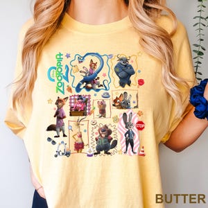 May include: A pale yellow t-shirt featuring a colorful graphic print of various characters from the movie Zootopia. The design includes multiple framed scenes, small icons like stars and paw prints, and the Zootopia logo in bright green text.