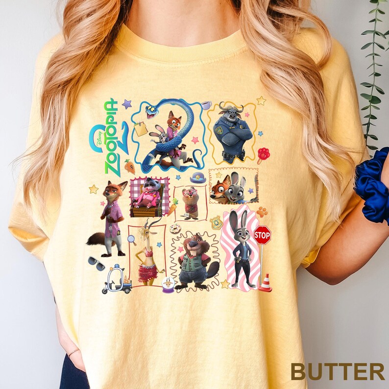 May include: A pale yellow t-shirt featuring a colorful graphic print of various characters from the movie Zootopia. The design includes multiple framed scenes, small icons like stars and paw prints, and the Zootopia logo in bright green text.