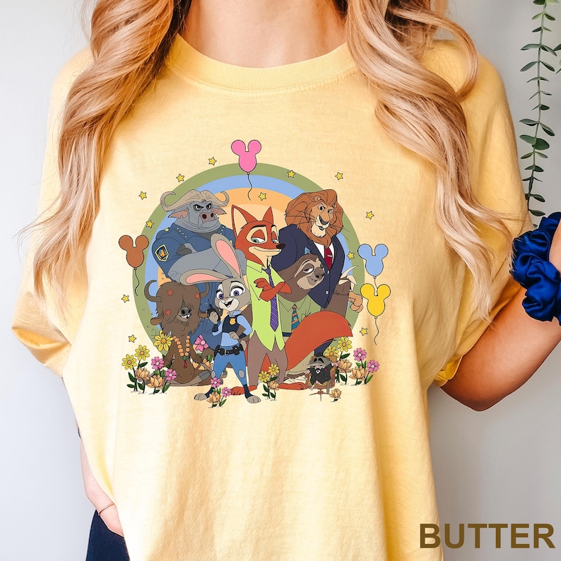 K&ouml;nnte beinhalten: Hellgelbes T-Shirt mit einer bunten Cartoon-Grafik von Zootopia-Charakteren. Das Design zeigt Nick Wilde, Judy Hopps und andere Charaktere, umgeben von Blumen, Luftballons und einem Regenbogen. Das Wort "BUTTER" ist unten rechts aufgedruckt.