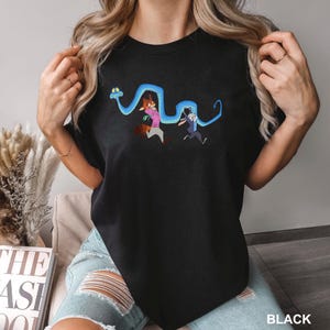 Op de afbeelding: Zwart T-shirt met een cartoon illustratie van twee personages die wegrennen van een blauwe slang. De personages zijn een vos in een roze shirt en een konijn in een blauw pak. Het woord "BLACK" staat onderaan.