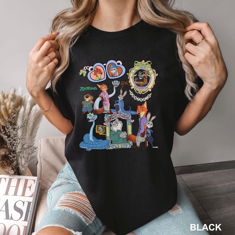 May include: A black t-shirt featuring a colorful graphic print of various Zootopia characters, including Nick Wilde and Judy Hopps, arranged in a collage with ornate frames, hearts, and the Zootopia logo text printed in green and white.