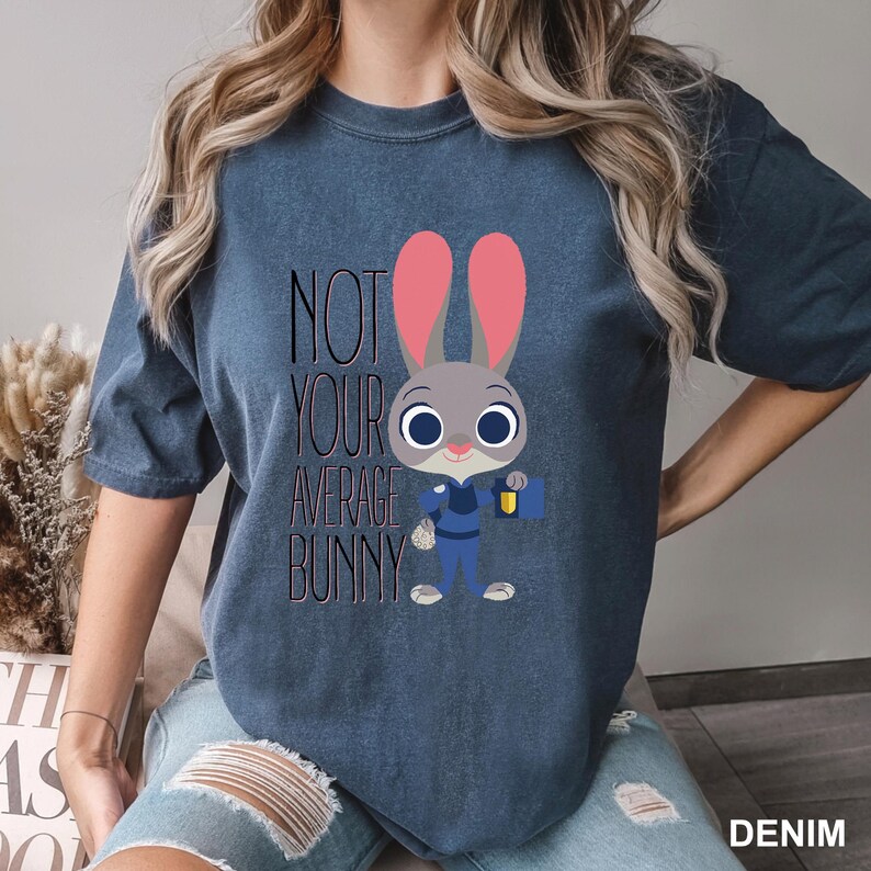 May include: Denim blue t-shirt featuring a cartoon bunny character with pink ears and a police uniform. The text "NOT YOUR AVERAGE BUNNY" is printed above the character. The shirt is a casual style.