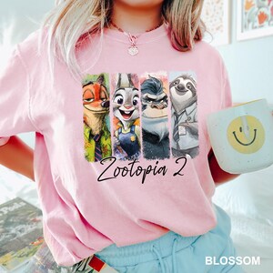 Peut inclure: Sweat-shirt rose clair avec un imprim&eacute; color&eacute; des personnages de Zootopia 2. Le design comprend Nick Wilde, Judy Hopps et d'autres personnages. Une tasse blanche avec un smiley jaune est tenue &agrave; la main.