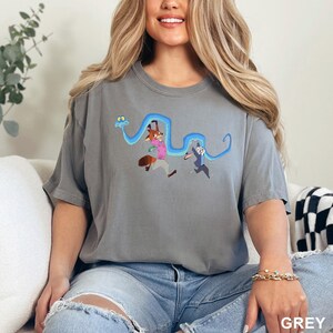 Op de afbeelding: Grijze t-shirt met een cartoonontwerp van een vos, een konijn en een blauwe slang. De vos draagt een roze shirt en het konijn een blauw shirt. Het woord "GREY" staat rechtsonder afgedrukt.