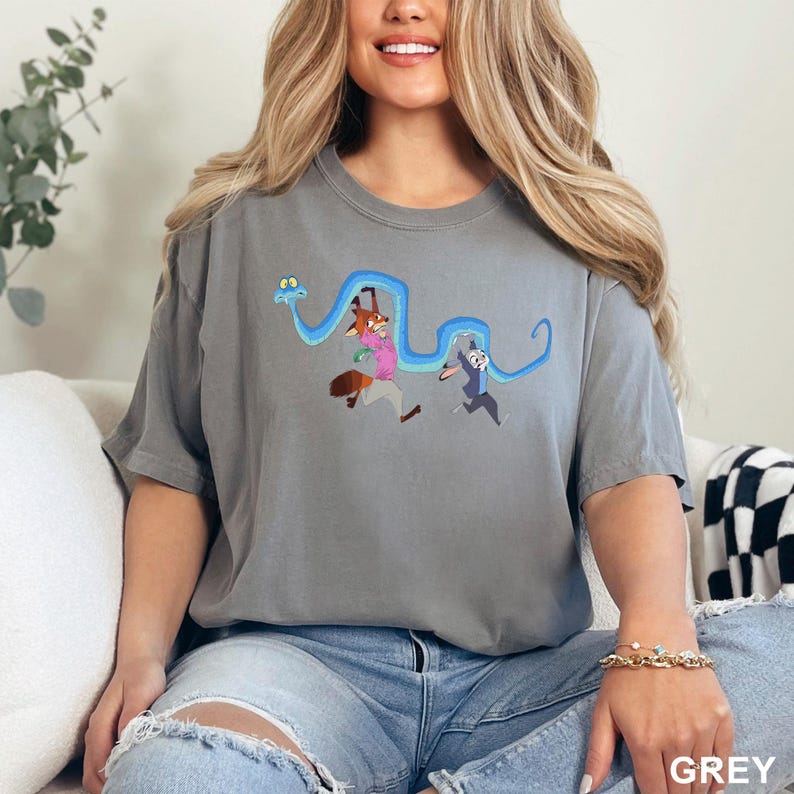 Op de afbeelding: Grijze t-shirt met een cartoonontwerp van een vos, een konijn en een blauwe slang. De vos draagt een roze shirt en het konijn een blauw shirt. Het woord "GREY" staat rechtsonder afgedrukt.