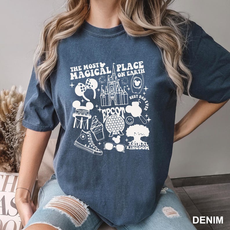Puede incluir: Camiseta azul denim con un dise&ntilde;o gr&aacute;fico blanco con el texto "THE MOST MAGICAL PLACE ON EARTH" y varios iconos tem&aacute;ticos de Disney, incluyendo un castillo, orejas de Mickey Mouse y la esfera de EPCOT.